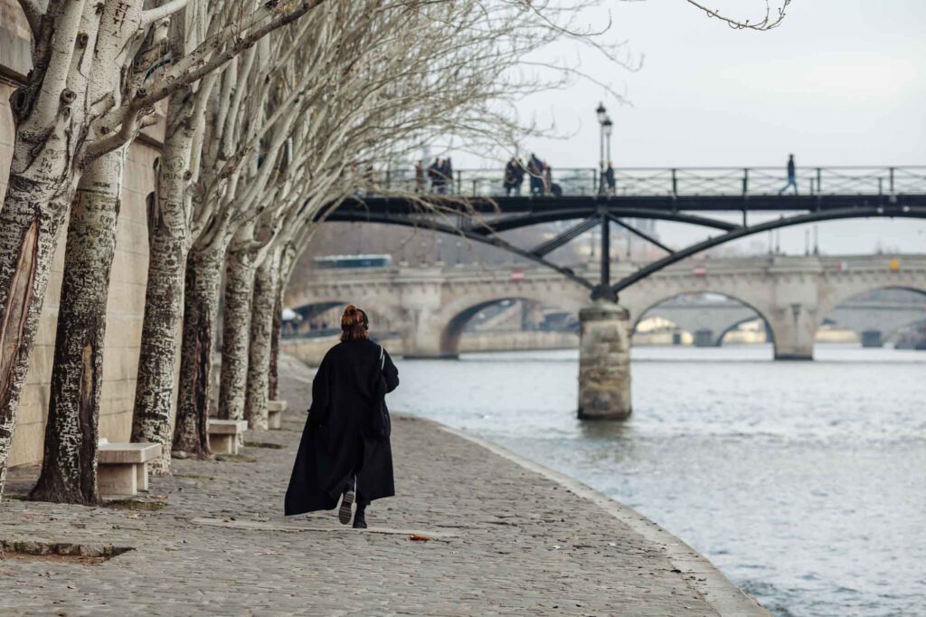 A woman in a black coat is walking along the Seine embankment in Paris