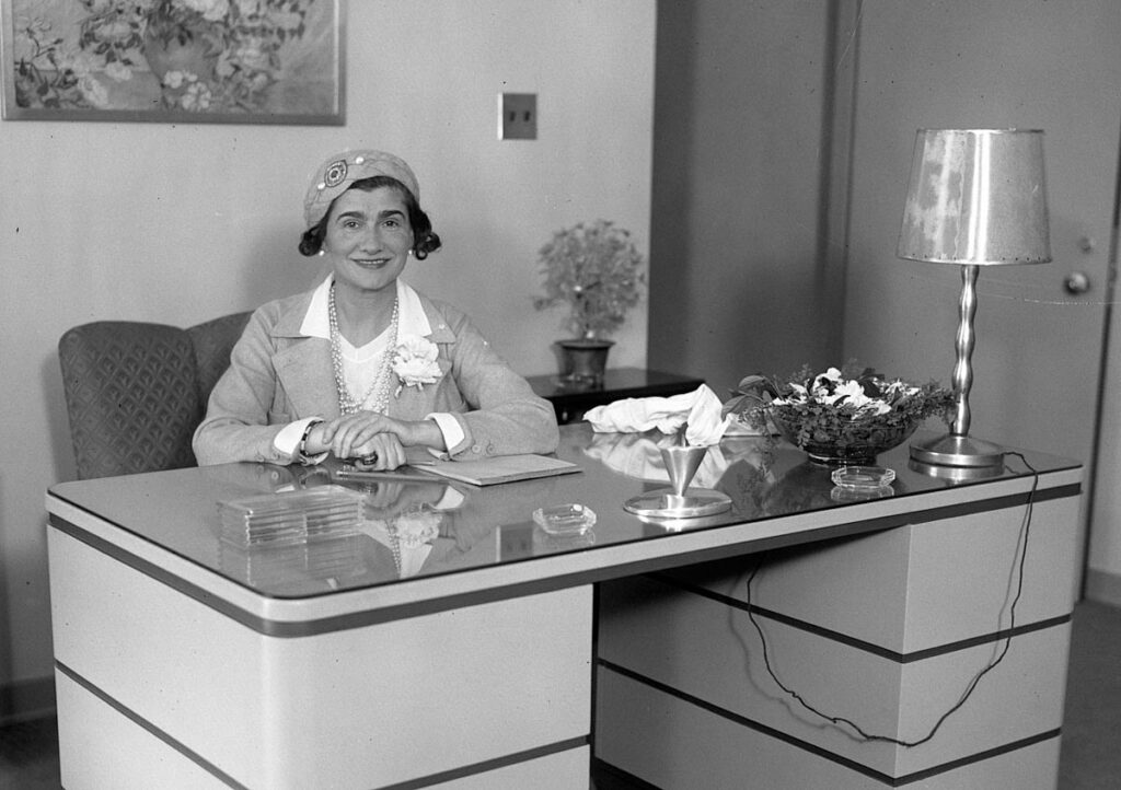 The Legendary Coco Chanel sitting behind a desk